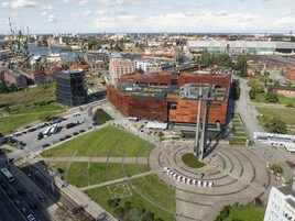 Egy városképet ábrázoló légifelvétel, amelyen egy feltűnő épület látható, egyedi tetőzetével, melyet utak, zöld területek és más épületek vesznek körül.