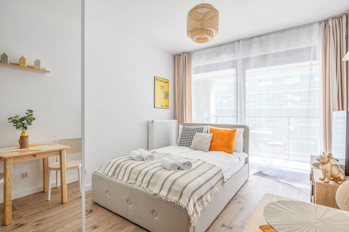 A modern bedroom with a large bed, a bedside table, a chair, a window with sheer curtains, and a small shelf with decorative items.
