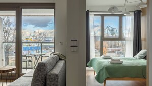 A modern room with a sofa, a bed with a green bedspread, and a view of a construction site through large windows. -  LILY APART - Rooftop Terrace Apartments by the Shipyard (Gdansk)