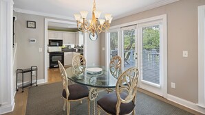 A dining area with a glass table, four chairs, a chandelier, a kitchen in the background, and large windows with blinds. - 28 Pelican Bay! Wild Dunes Family Vacation! Community Pool, Golf Views, and Close to Beach! (Isle Of Palms)