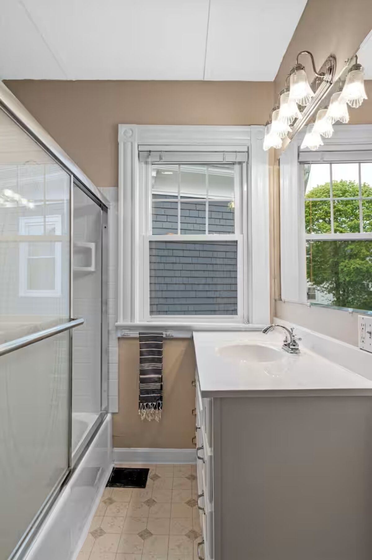 A modern bathroom with a glass shower enclosure, a window with blinds, a sink with a mirror, and a towel rack.