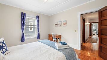 Quarto com cama, escrivaninha e cadeira. Cortinas na janela e porta que dá para outro cômodo.