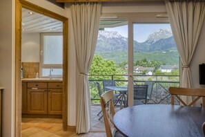A room with a view of mountains, a dining area with a table and chairs, and a kitchen with wooden cabinets and a window. - Roseaux du château (Duingt)