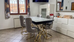 A modern dining area with a glass-top table, cushioned chairs, a television, and a kitchen counter with a coffee maker and a red first aid box. - B&B La Fenice Rome - Fiumicino. Close to the center of Rome and the airport. (Lazio)