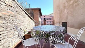 Une table ronde recouverte d’une nappe à motifs, entourée de chaises en métal blanc, placée devant un mur de pierre et un bâtiment à la façade rose.