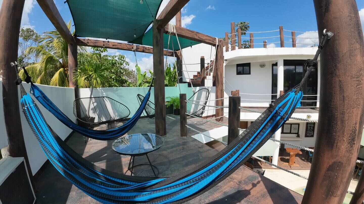 A hammock chair and a small table on a wooden deck with a view of a white building and palm trees.