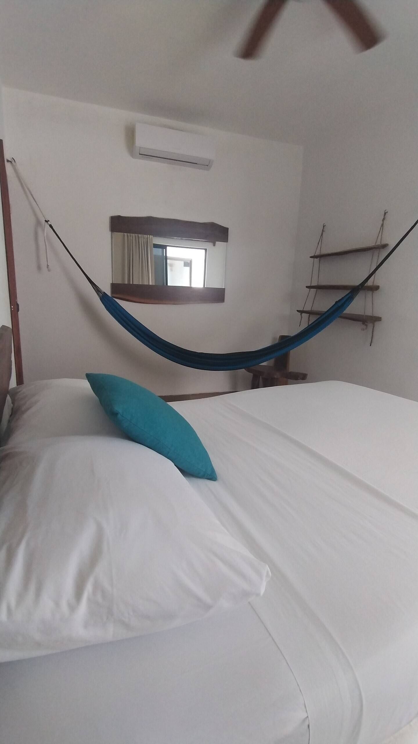 Una cama con sábanas blancas y almohada azul, una hamaca, una escalera de madera y un espejo en la pared.