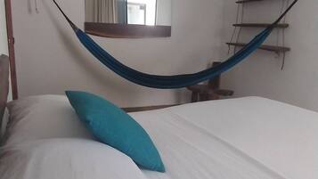 Una cama con sábanas blancas y almohada azul, una hamaca, una escalera de madera y un espejo en la pared.