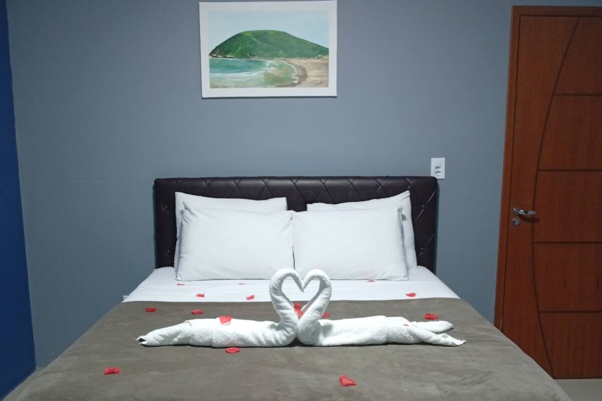 A bed with a brown headboard, white pillows, and a heart-shaped towel arrangement. A framed picture of a beach is on the wall.