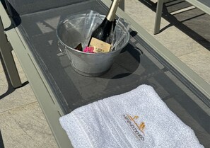 Una mesa metálica con un cubo metálico que contiene una botella y una tarjeta, y una toalla blanca con texto.