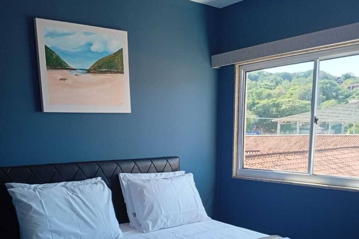 A hotel room with a bed, a painting, and a window with a view of buildings.