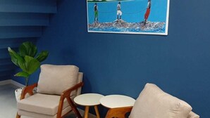 A room with blue walls, a painting of people by the sea, and two beige armchairs with small white tables. - Pousada Menino do Rio (Buzios)