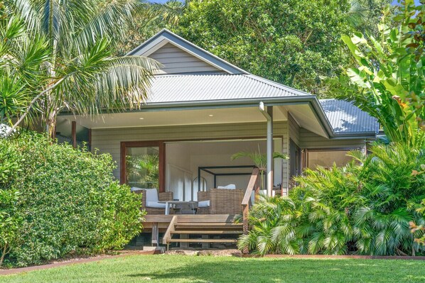 Relax on the porch with a wicker chair and small table, surrounded by lush vegetation and a well-manicured lawn.