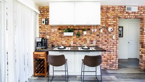 Une cuisine avec un mur en briques, deux tabourets de bar, un meuble à bouteilles et une horloge au mur.