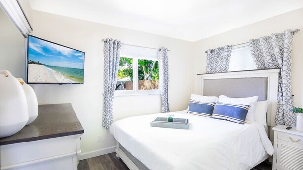 Une chambre à coucher avec un lit, un téléviseur fixé au mur, une fenêtre avec des rideaux et une table de chevet.