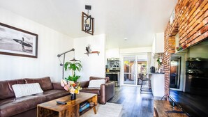 Un salon moderne avec un canapé en cuir marron, une table basse en bois et un téléviseur. Il y a également un coin cuisine avec un mur en briques et une porte-fenêtre coulissante donnant sur une terrasse.