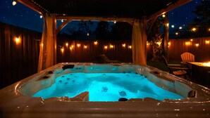 Un bain à remous sous une pergola en bois, illuminé de guirlandes lumineuses, entouré d’une clôture en bois et de chaises.