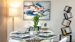 A modern dining area with a round glass table, white cushioned chairs, a contemporary painting above, and a bookshelf with books. - ` Velvet Bloom ` unique location 3mil to Downtown (Naples)