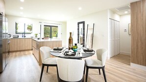 A modern kitchen with a dining area, featuring a central island, white chairs, and a large window providing natural light. - ` The Black Vanilla ` Luxe Retreat Sauna, Cold Plunge & Pure Relaxation (Naples)