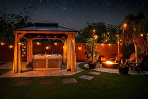 A gazebo with a hot tub, outdoor seating, and a fire pit, surrounded by a wooden fence and potted plants.