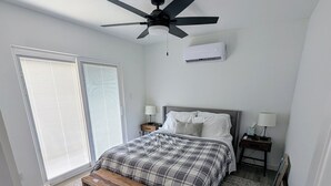 A bedroom with a bed, a ceiling fan, a nightstand, a bedside table, a bench, a window with blinds, and an air conditioning unit. - Wonderful Pondview Cottage in College Station (College Station)