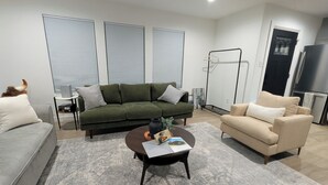 A modern living room with a green sofa, a beige armchair, and a grey sofa. There is a coffee table with a book and a vase. The room has large windows with blinds, a black door, and a black metal rack. - Wonderful Pondview Cottage in College Station (College Station)