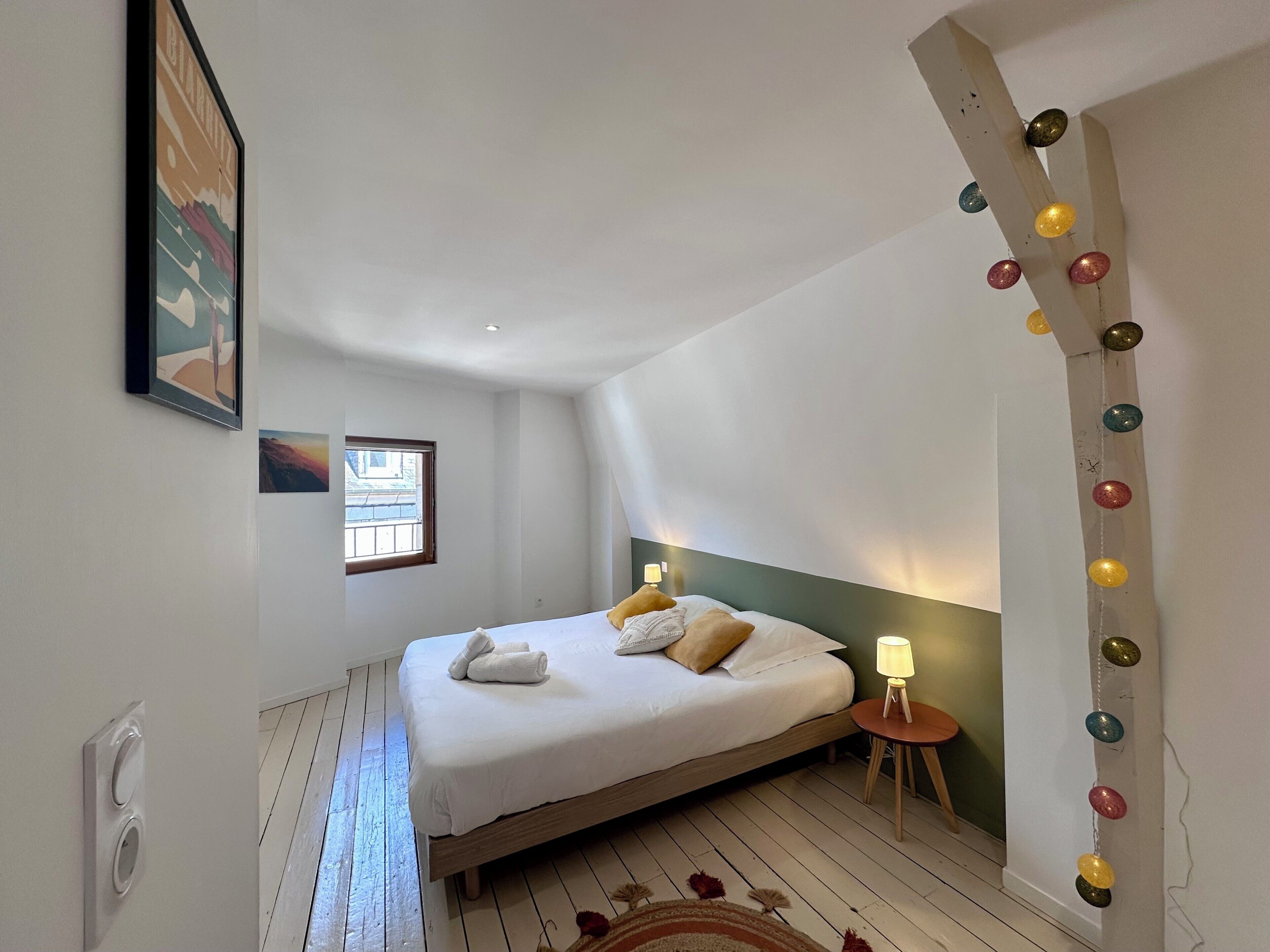 Ein Schlafzimmer mit einem Bett, einer Nachttischlampe, einem Fenster und einem an der Wand hängenden Bild.