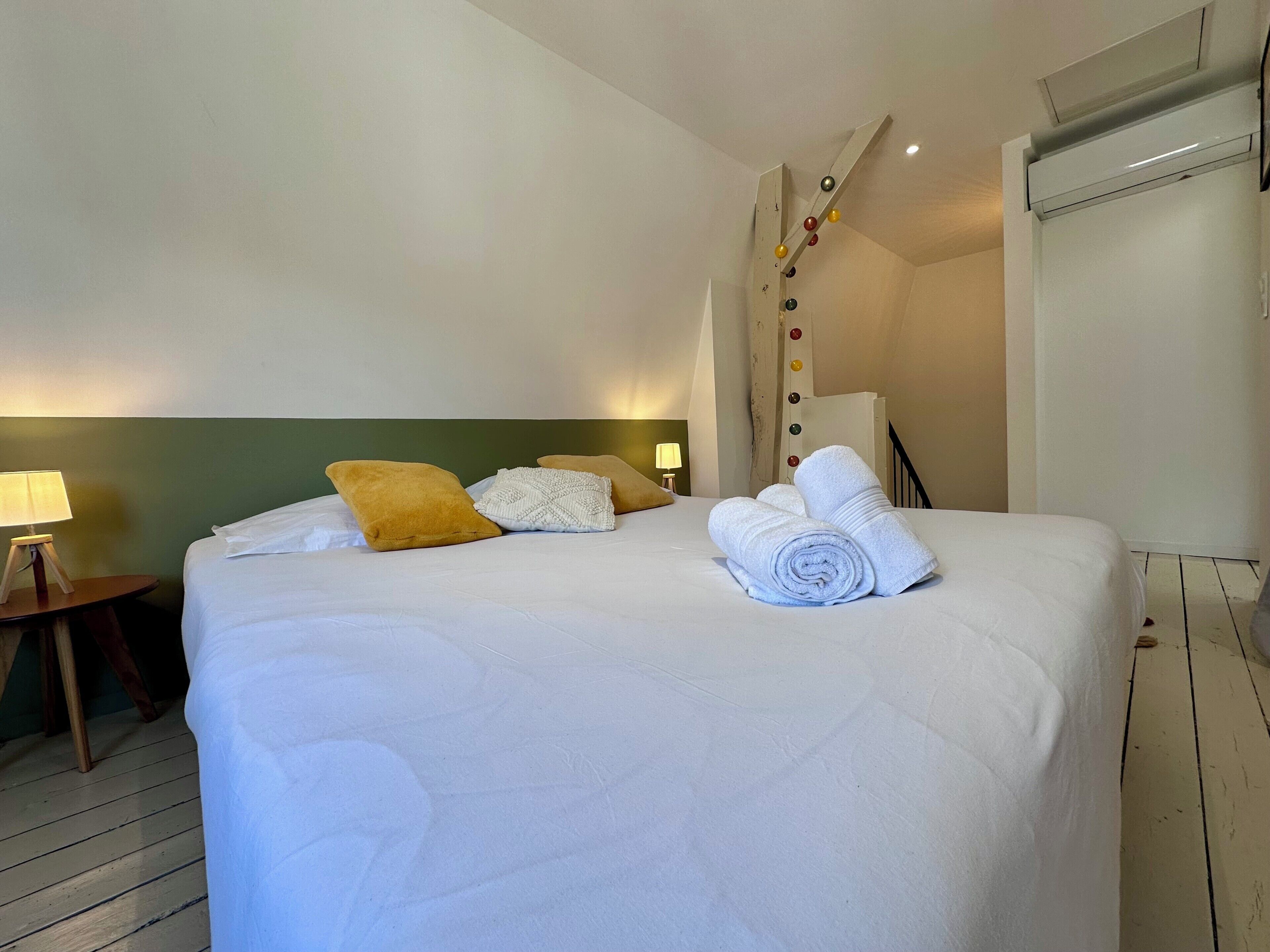 Een net opgemaakt bed met witte en gele kussens, een opgerolde witte handdoek en een wandlamp met gekleurde knoppen.