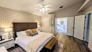 Un dormitorio con cama, mesitas de noche, un banco, un ventilador de techo y un baño visible a través de una puerta abierta.