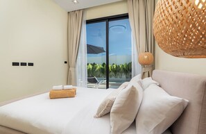 A hotel room with a large bed, a view of greenery through a sliding door, and a wicker lamp.