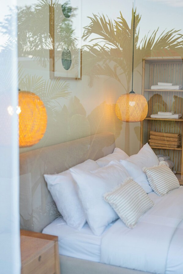 A bed with white bedding and striped pillows, a woven pendant light, and a shelf with folded towels.