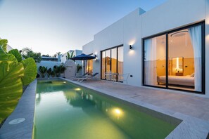 Un hôtel moderne doté d’une piscine, de chaises longues et d’une chambre bien éclairée, visible à travers de grandes portes coulissantes.