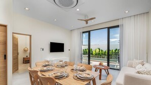 A modern living room with a dining area, a flat-screen TV mounted on the wall, and a large sliding glass door leading to a balcony with a view of greenery. - Staylar Pool Villa - Bangtao, Phuket C1 (Phuket, Bangtao)