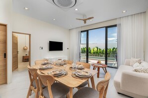 Un salon moderne comprenant un coin repas, un téléviseur à écran plat fixé au mur et une grande porte-fenêtre coulissante donnant sur un balcon avec vue sur la verdure.