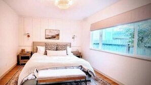 A bedroom with a large bed, bedside tables, a nightstand, a window with a view, and a ceiling light fixture. - Downtown Designer Duplex close to Beach! Spa Tub, Sunroom, Game Room, EV & more! (Long Beach)
