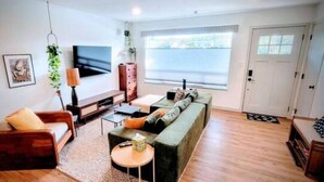 A modern living room with a sofa, armchair, TV, and a large window. - Downtown Designer Duplex close to Beach! Spa Tub, Sunroom, Game Room, EV & more! (Long Beach)
