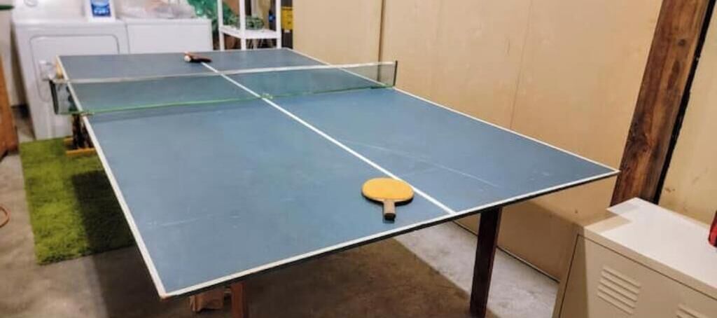 Ein Tischtennistisch mit Schläger in der Mitte, eine Waschmaschine und ein Schrank im Hintergrund.