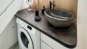 A modern bathroom with a built-in washing machine, a stone sink, and a round mirror with built-in lighting.