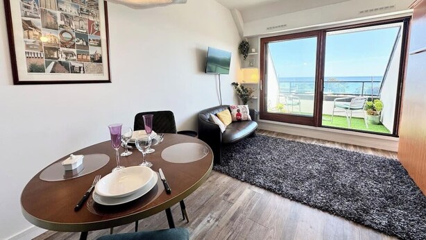 A modern living room with a dining table set for two, a black leather sofa, and a large window offering a view of the sea.