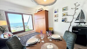 A dining area with a table set for four, a sofa, a wooden cabinet, and a view of the sea through large windows.