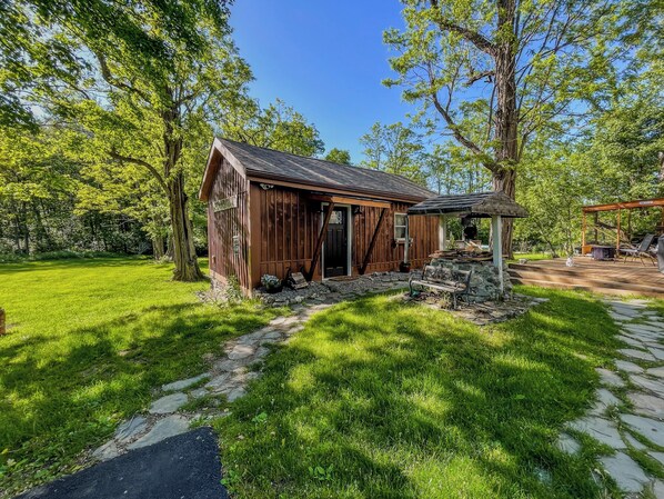 A rustic wooden cabin with a porch, surrounded by lush greenery and a stone pathway.