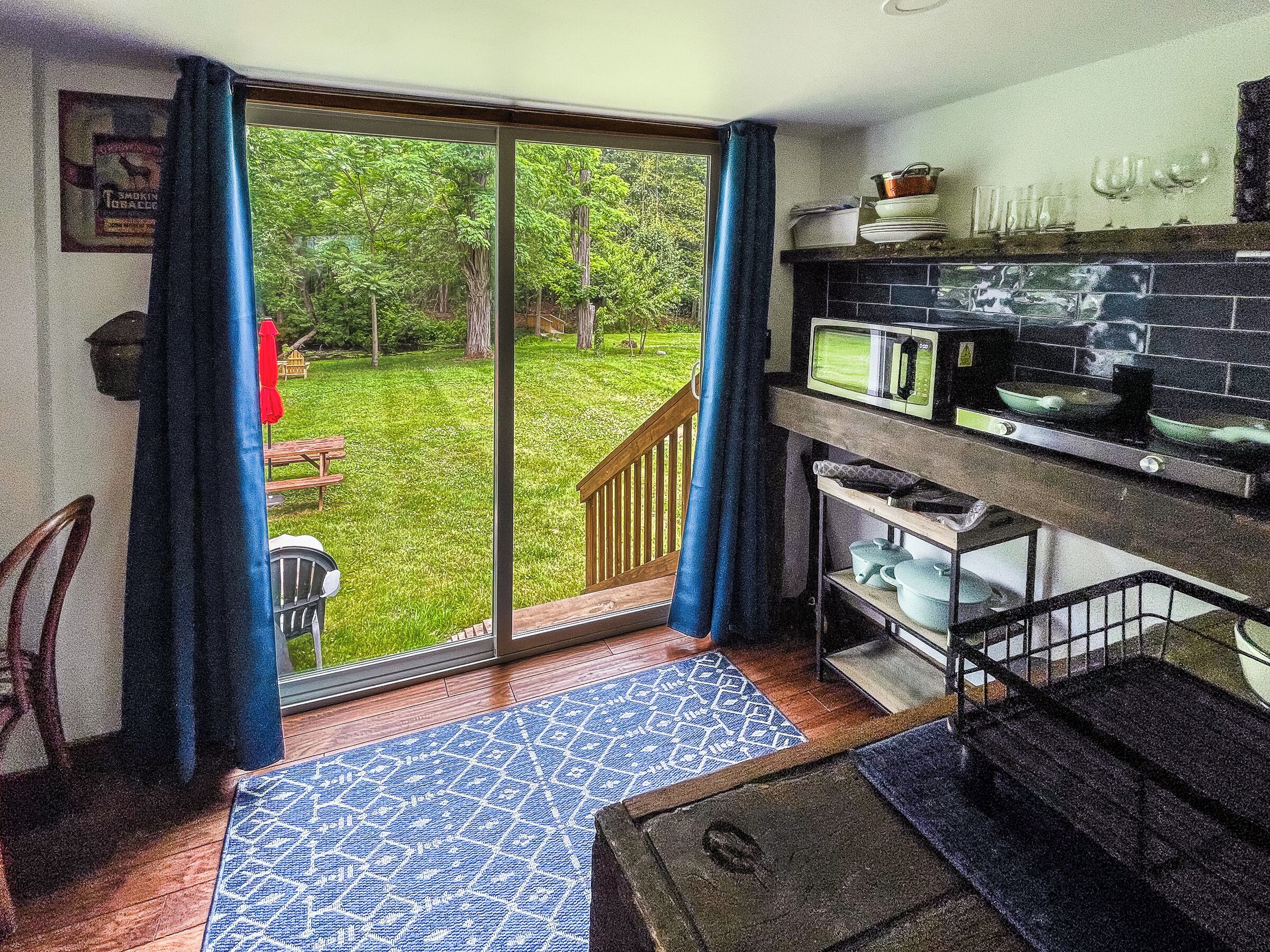 Une cuisine équipée d’un four à micro-ondes, d’une cuisinière et d’étagères, une porte-fenêtre coulissante donnant sur un jardin, et un tapis bleu à motifs.
