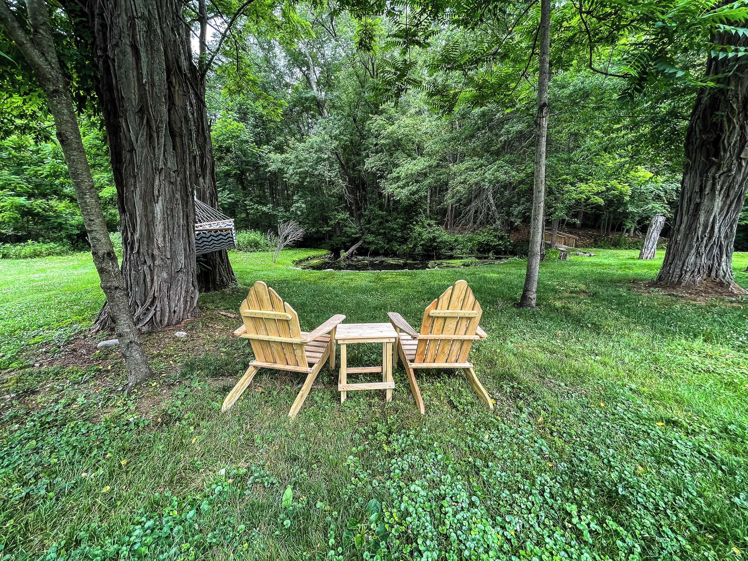 A wooden table and two Adirondack chairs in a grassy area surrounded by trees.