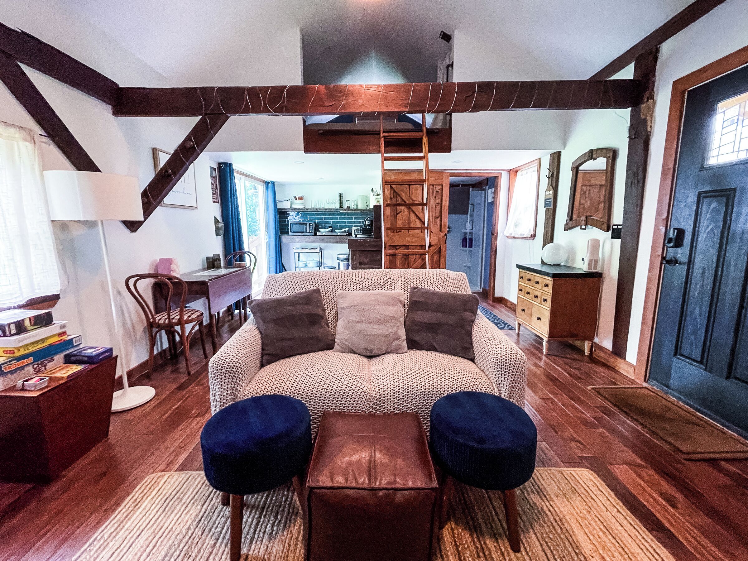 A cozy room with a patterned sofa, two ottomans, a wooden ladder, a dining area, and a kitchen in the background.