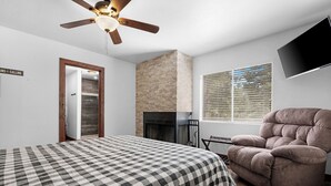 Ein Schlafzimmer mit kariertem Bettwäsche, Kamin, Deckenventilator, Wandfernseher und einem Fenster mit Jalousien.