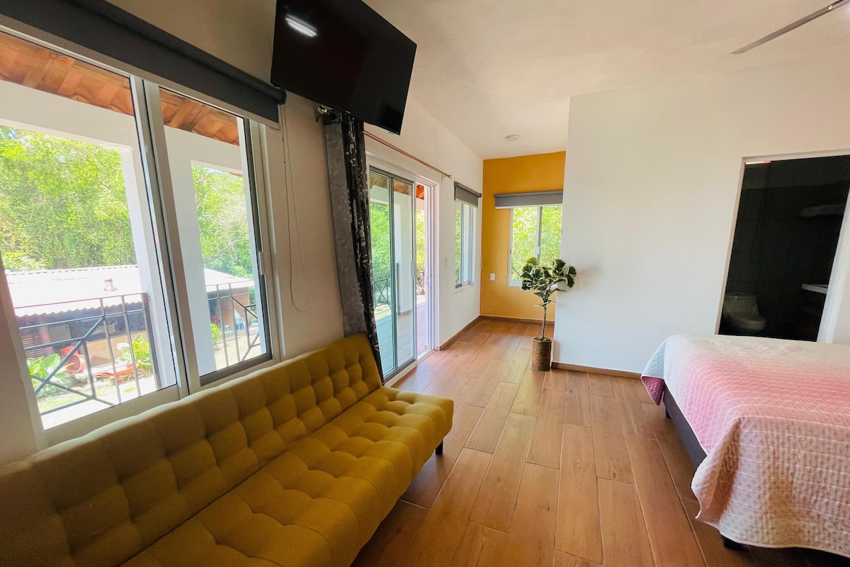 A room with a yellow sofa, a bed with a pink blanket, a TV mounted on the wall, and large windows with a view of greenery outside.