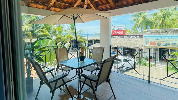 A covered outdoor seating area with a table and chairs, a potted plant, and a view of a beach and palm trees. - Bungalows Garza Blanca a Pie de Playa (San Blas)