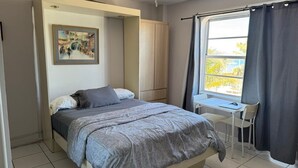 A bedroom with a bed, a desk, and a chair. There is a window with curtains. - Charming studio in sunny Miami Beach with ocean view! (Miami Beach)