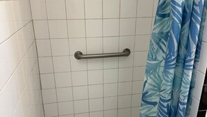A bathroom with white tiled walls, a grab bar, and a shower curtain with a leaf pattern. - Charming studio in sunny Miami Beach with ocean view! (Miami Beach)