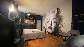 A room with a large mural, a bed with a white cover, a small table, and a chair.